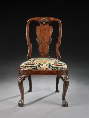 A single chair of dark brown colour, with a rich golden dark brown centre back splat. The seat is upholstered with a needlework depicting a woman dressed in early 18th century period blue dress with surrounding foliage and floral pattern typical of the time in colours of black, ochre, red and greens . 